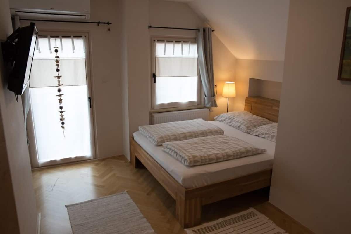 A bedroom with a wooden bed, two windows with curtains, a radiator, and a lamp.