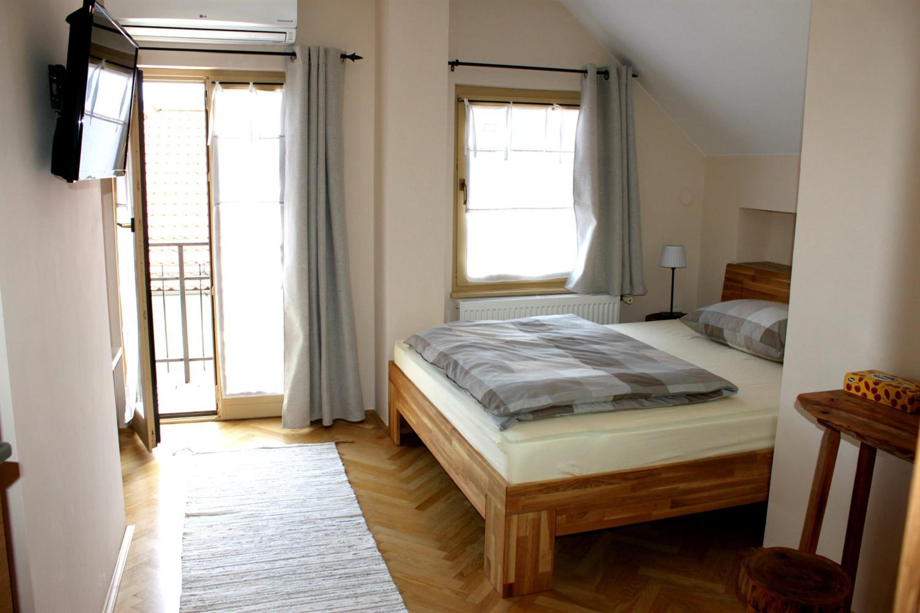 A bedroom with a wooden bed, a window with curtains, a radiator, and a small table with a lamp.