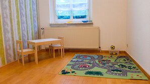 Une salle de jeux pour enfants avec une table et des chaises en bois, un tapis de jeu coloré, une fenêtre avec des stores et un radiateur.