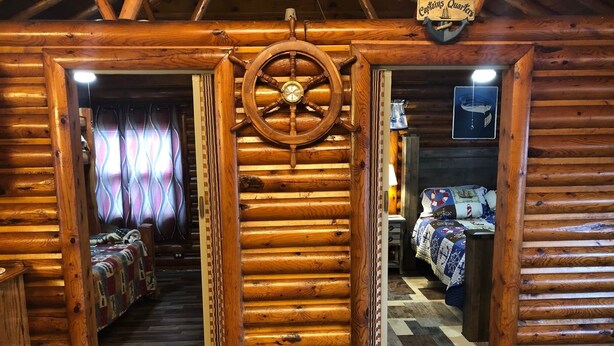 Interior de una cabaña de troncos con una puerta de madera central que da a un dormitorio, una cama con colcha y una mesita con lámpara.