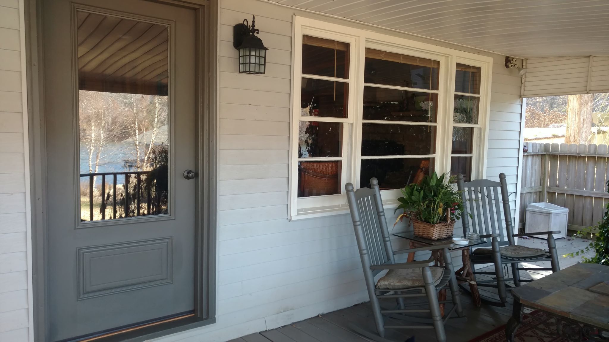 A porch with a gray door, a white window with multiple panes, and two wooden chairs with cushions.