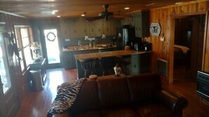 A cozy cabin interior with a wooden ceiling, a kitchen area, a dining table, and a leather couch.