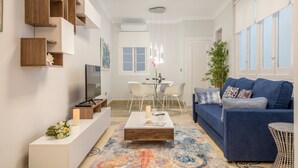 Een moderne woonkamer met een blauwe bank, een bijzettafeltje, een televisie en een eethoek met een witte tafel en stoelen.