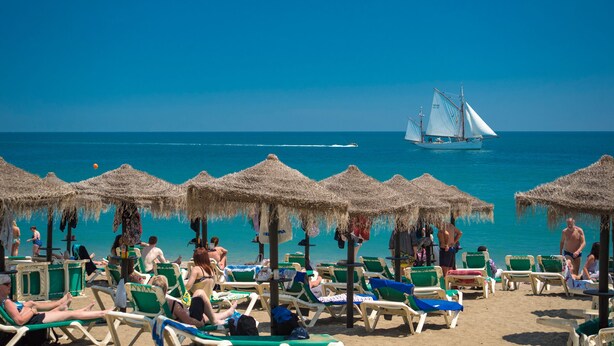 Een strand met rijen ligbedden onder rieten parasols, een zeilboot in de verte en helderblauw water.