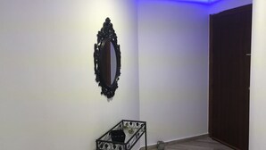 Una habitación con una puerta de madera oscura, una pared blanca con un espejo decorativo y una pequeña mesa de metal negro con superficie de vidrio.