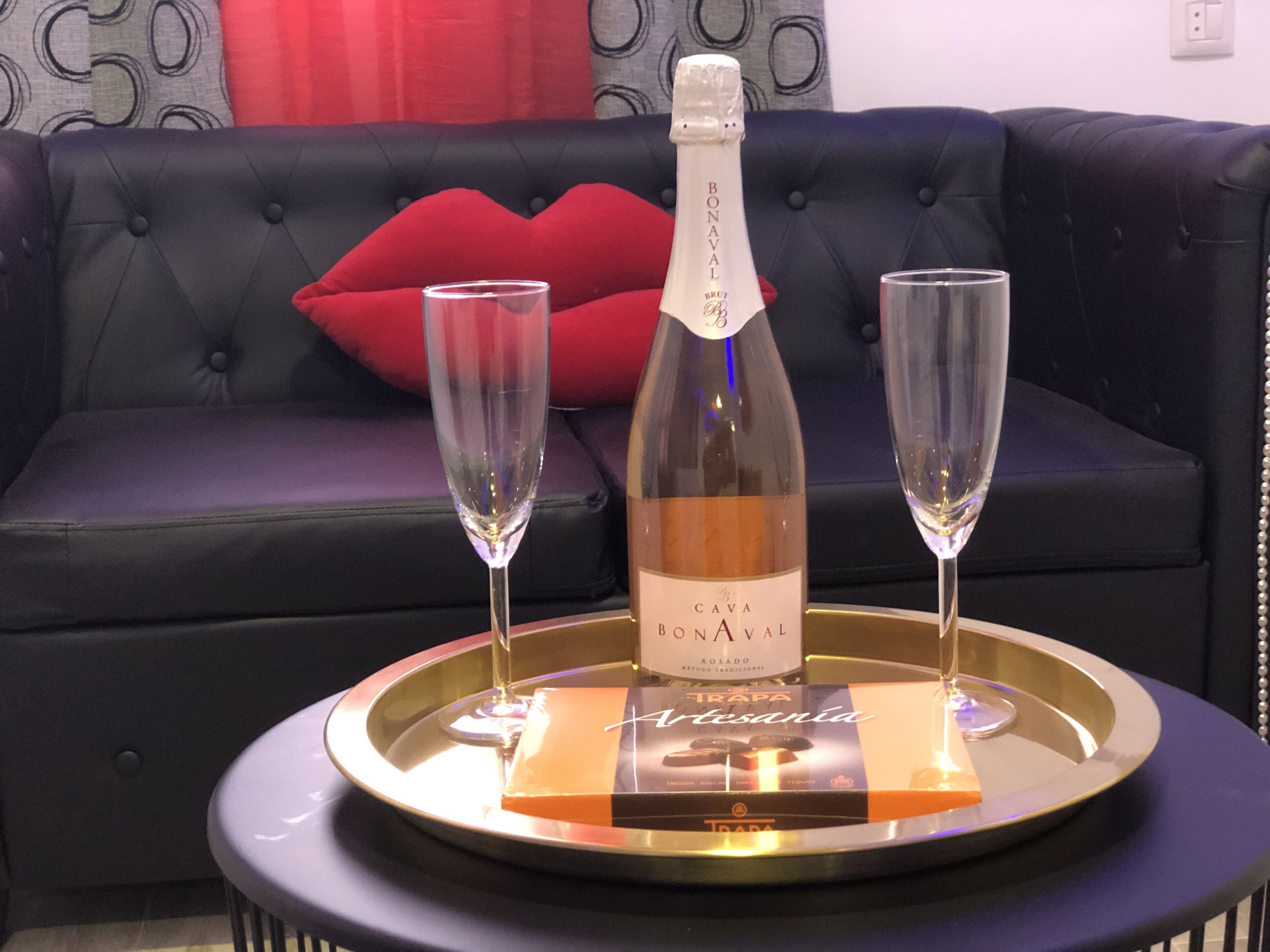 A bottle of Cava Bonaval on a tray with two glasses, set on a table in front of a dark blue sofa with a red pillow.