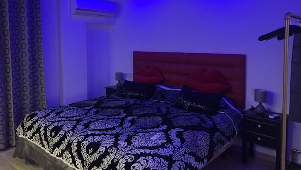 Un dormitorio con una lámpara circular en el techo, una cama con ropa de cama estampada, un cabecero rojo y una mesita de noche con lámpara.
