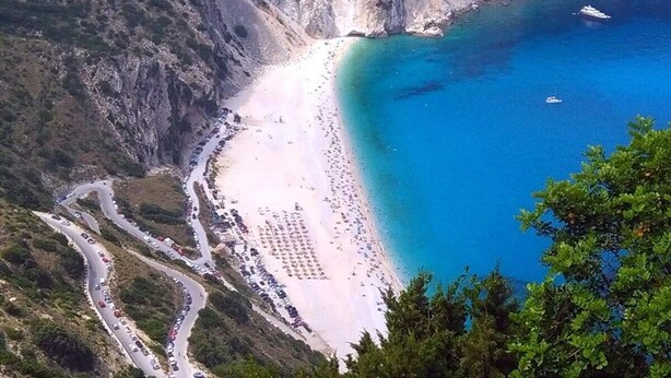 Una carretera costera que lleva a una playa con mar azul cristalino, rodeada de acantilados.