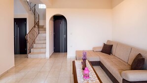 Una sala de estar con un sofá color crema, una mesa de centro con un florero y un farol rosa, una escalera con barandilla negra y una puerta negra.