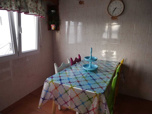 A dining area with a table covered by a colorful tablecloth, a clock on the wall, and a window with a floral curtain.