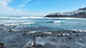 Une ville côtière aux bâtiments blancs qui bordent le littoral, avec une plage rocheuse et des vagues qui se brisent sur le rivage.