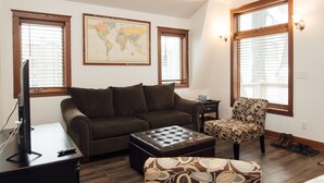 A living room with a brown sofa, a patterned armchair, a coffee table, and a television on a stand. There are two windows with blinds, one map on the wall, and a lamp on the right side. - Kingfield, Minneapolis; Sleeps 6; New Construction, 3 Miles from Downtown (Minneapolis)