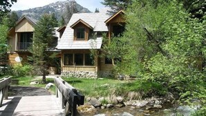 A wooden cabin with a stone foundation, surrounded by lush greenery and a small stream.