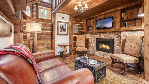 A cozy log cabin interior with a leather sofa, two armchairs, a fireplace, a television, and a staircase.