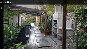 Eine überdachte Terrasse mit Stein-Barbecue, Gartenmöbeln und einem Gartenbereich.