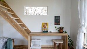 Una habitación pequeña con un escritorio de madera, una silla blanca, un taburete blanco, una escalera, una ventana y una planta en maceta.