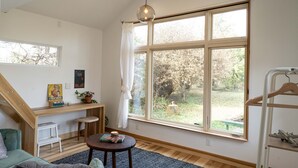 Una habitación luminosa con un amplio ventanal, un escritorio de madera, un taburete pequeño, una mesa redonda y una lámpara colgante.