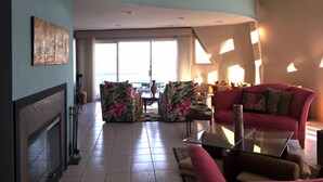 Un salón espacioso con chimenea, un sofá rojo, una mesa de centro de cristal y vistas al océano a través de amplias ventanas.