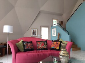 Uma sala de estar com um sofá vermelho, almofadas coloridas, um abajur e uma escada que leva a um andar superior.