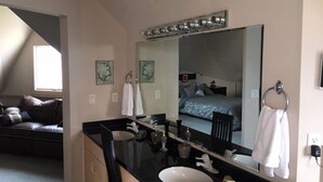 Un baño moderno con un espejo grande, un lavamanos y un inodoro. Se ve una cama al fondo.