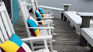 Una hilera de sillas Adirondack blancas con cojines de colores en una terraza de madera junto al agua.