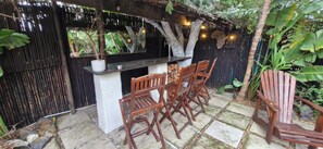 Un área de estar al aire libre con una mesa y sillas de madera, una planta en maceta y un fondo de árboles y follaje.