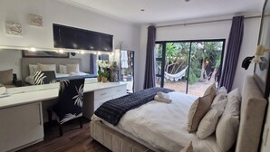 A bedroom with a bed, desk, chair, and a large window with a view of a hammock and greenery.