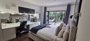 A bedroom with a bed, desk, chair, and a large window with a view of a hammock and greenery.