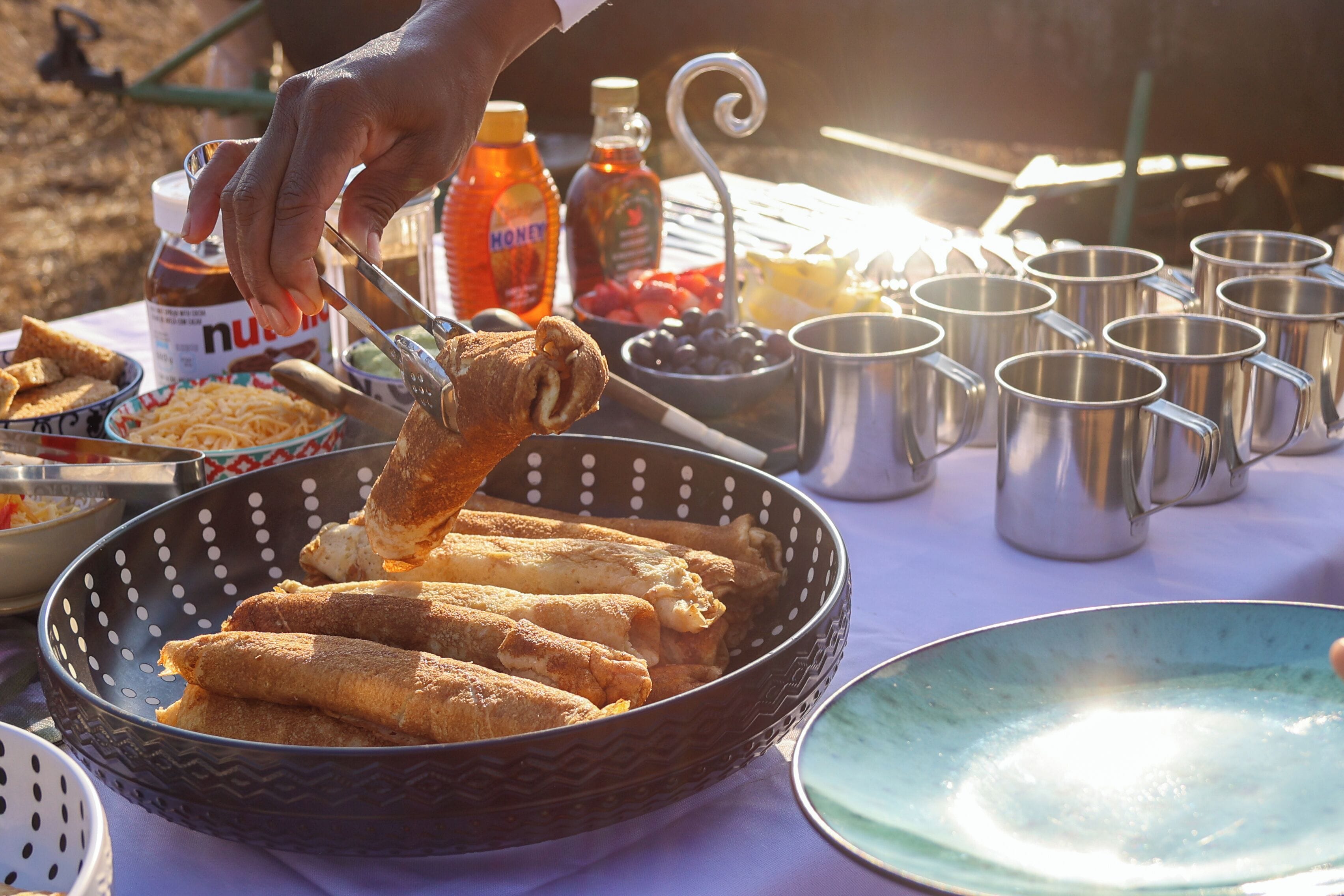 A table with food items, including a basket of fried rolls, a bowl of noodles, and several metal cups.