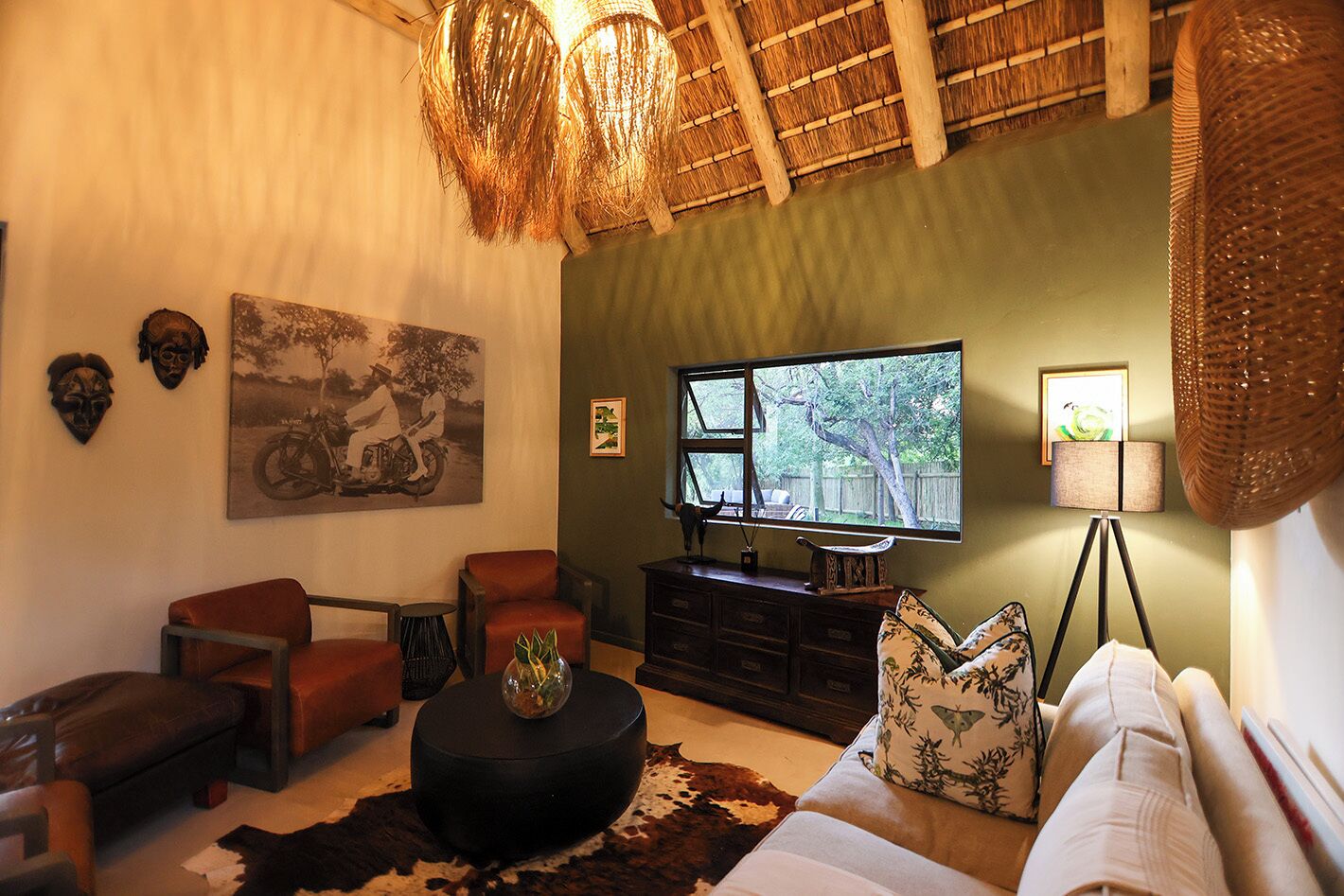 A living room with a sofa, two armchairs, a coffee table, a TV, and a dining table. There are decorative masks on the wall and a large window with a view of trees.