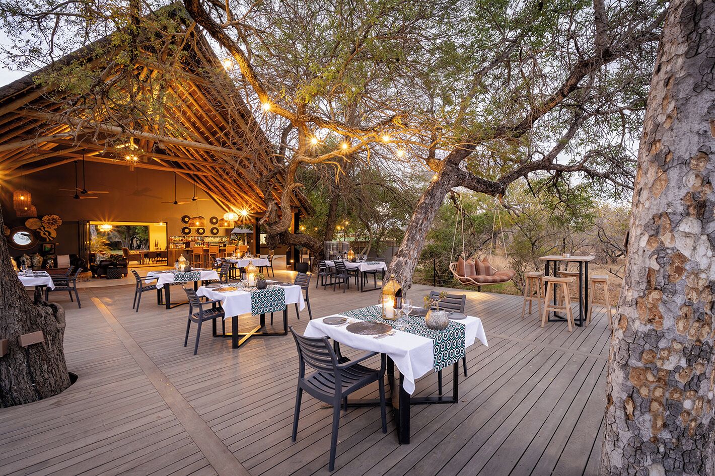 Un área de comedor al aire libre con mesas dispuestas para los invitados, rodeada de árboles y una terraza de madera.