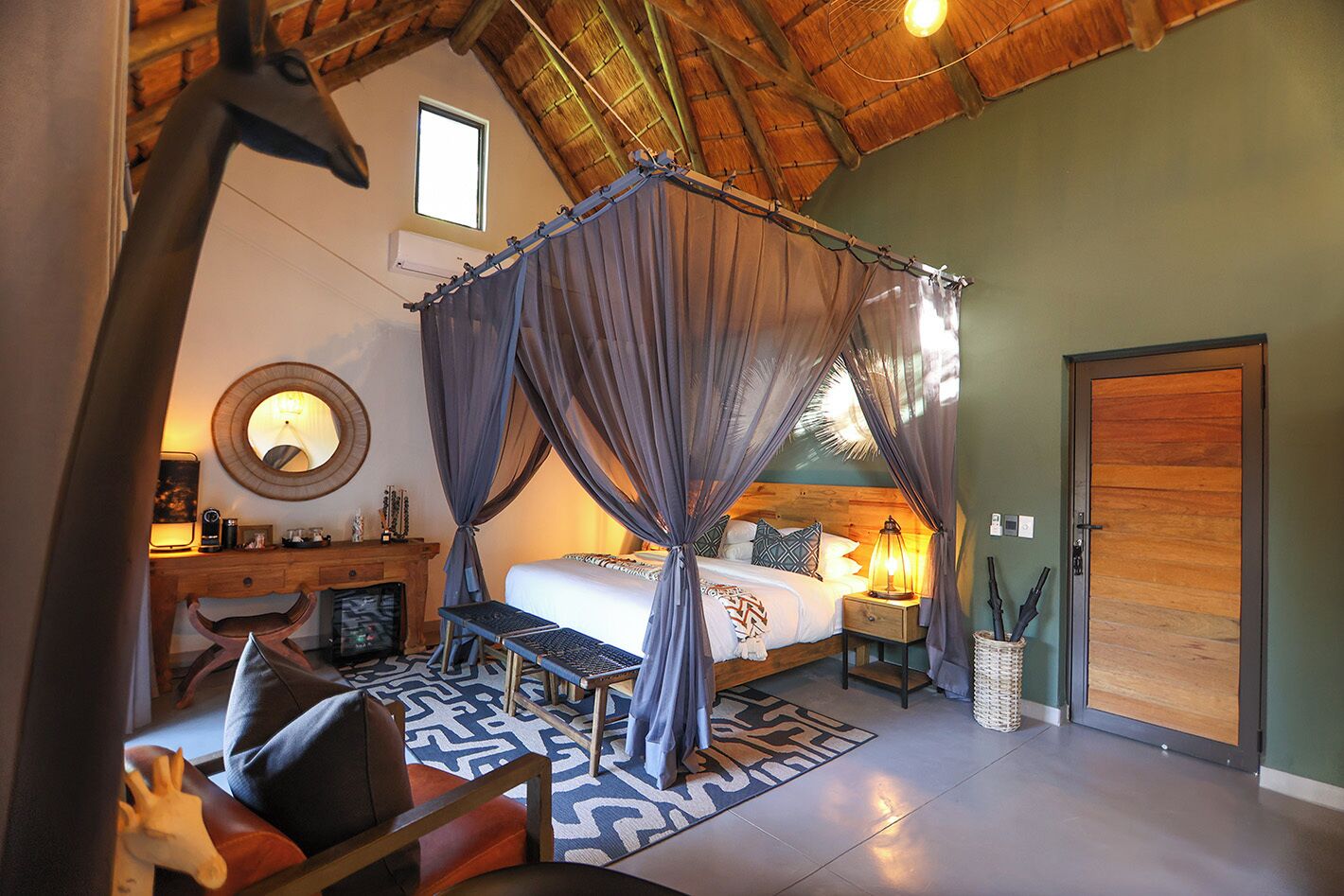 A bedroom with a canopy bed, a desk with a chair, a bedside table with lamps, and a large mirror on the wall.