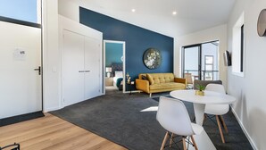 A modern living room with a sofa, a round table, and two chairs. There is a door leading to another room and a window with blinds. - Modern One Bedroom Apartment with Lake View (Queenstown)