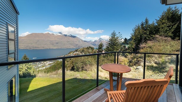 A balcony with a wooden table and chair, overlooking a lake and mountains. - Modern One Bedroom Apartment with Lake View (Queenstown)