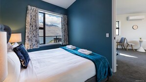 A hotel room with a bed, a window with curtains, a view of mountains, a small table with a plant, and a chair. - Modern One Bedroom Apartment with Lake View (Queenstown)