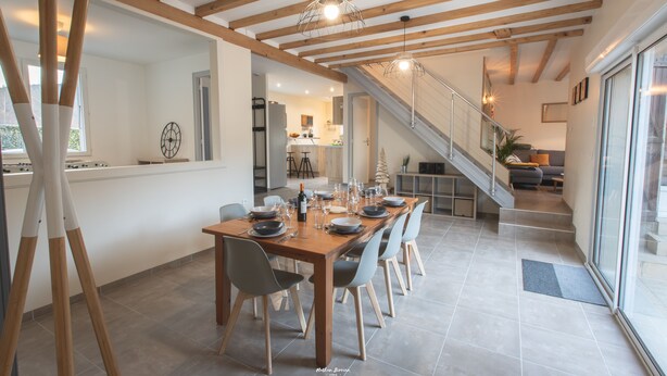 A dining area with a wooden table set for six, a staircase leading to an upper level, and a kitchen in the background.