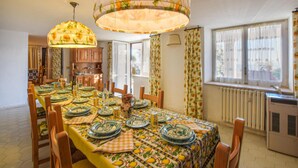 Comedor con una larga mesa dispuesta para seis comensales, rodeada de sillas de madera. Dos lámparas colgantes con motivos florales, y una ventana con cortinas.