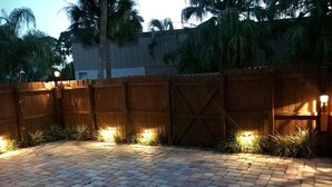 A paved outdoor area with a wooden fence and a gate, surrounded by plants and palm trees. - The Polynesian- 2 miles to Siesta Beach, Heated Pool, Furry Friends Stay Free (Sarasota)