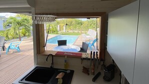 A compact kitchen with a built-in dishwasher, a window overlooking a pool area, and outdoor seating. - Villa close to all amenities and beach (Saint-François)