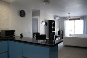 Una cocina moderna con encimera negra, microondas y un reloj en la pared. Hay una zona de comedor con una silla negra y un sofá blanco. Se ve un equipo de aire acondicionado montado en la pared, y también un ventilador de techo.