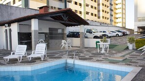 A swimming pool with white lounge chairs, a covered area with a tiled roof, and a parking lot with several cars. - SEA STIEP APARTMENT (Salvador)