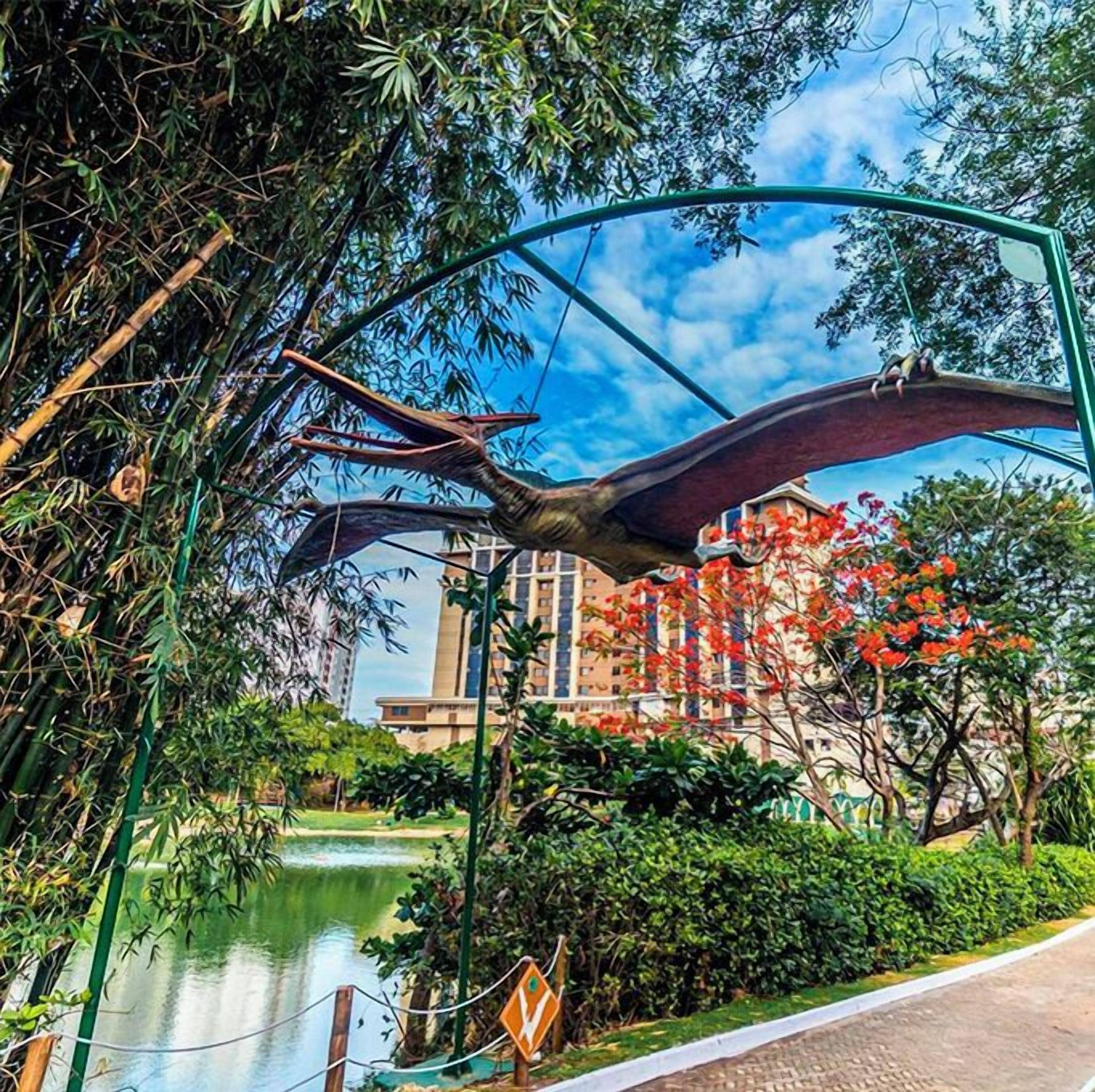 A large bird sculpture in a park with a pond, surrounded by greenery and buildings in the background.