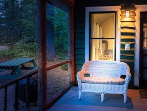 A wicker chair with a cushion on a wooden deck, a wooden bench, and a glass door leading to an interior space with a light fixture.