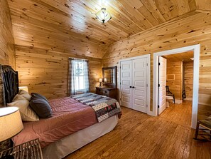 Quarto de cabana de madeira com cama, mesa de cabeceira, abajur, espelho e porta que dá para um banheiro.