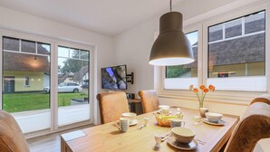 Un coin repas avec une table en bois dressée pour quatre personnes, un téléviseur fixé au mur et de grandes fenêtres donnant sur l’extérieur.