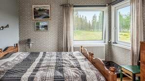 Et soveværelse med en stor seng, et træhovedgærde, en grøn stol, en radiator og et vindue med udsigt til en skov.