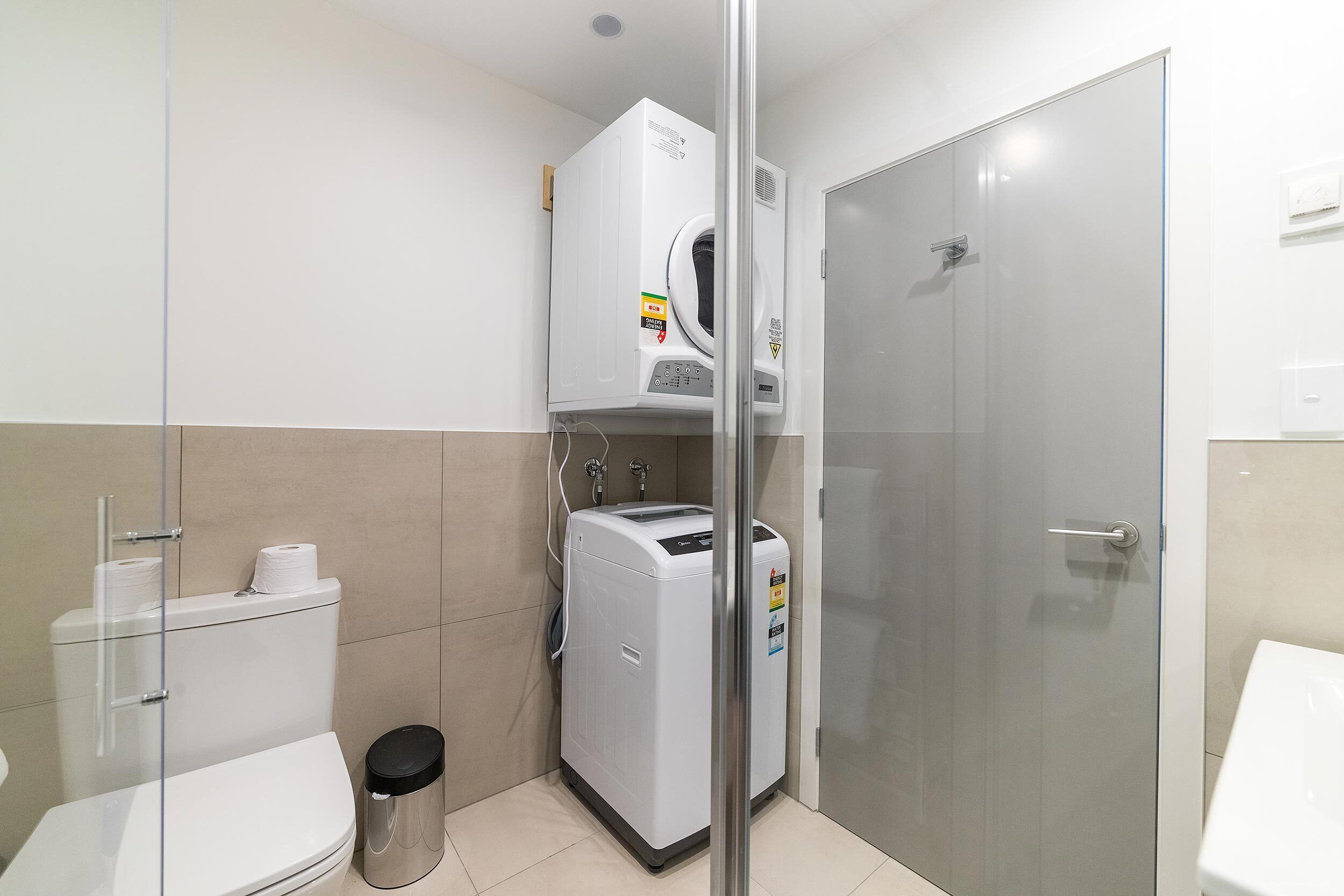 A compact bathroom with a toilet, a trash bin, a washing machine, and a glass shower enclosure.