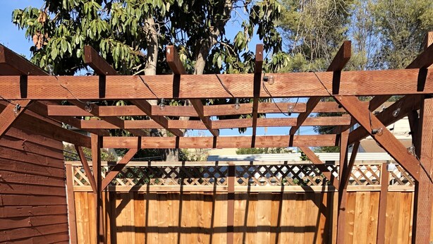 Un patio al aire libre con una pérgola de madera, un sofá beige con cojines rojos, dos sillas negras y una pequeña mesa.