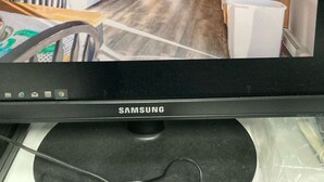Ein Samsung-Monitor zeigt eine Küche mit Holzschränken, einem Kühlschrank, einer Mikrowelle und einem Essbereich mit Tisch und Stühlen.