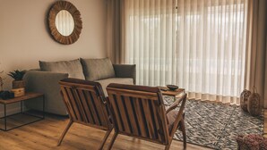 Un salón con un sofá, dos sillas de madera, una mesa de centro, un espejo redondo y un gran ventanal con cortinas translúcidas.